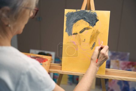Téléchargez les photos : Gray-haired woman paints a portrait with paints and a brush, drawing her hobby - en image libre de droit