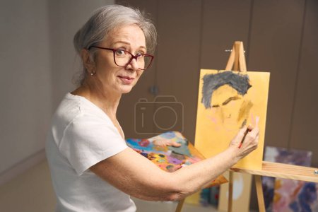 Foto de Talentosa anciana se para en un caballete con una paleta y un pincel, dibujando su hobby - Imagen libre de derechos
