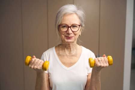 Téléchargez les photos : Heureuse retraitée faisant de l'exercice avec des haltères pour maintenir sa santé, elle est de bonne humeur - en image libre de droit