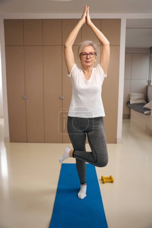Foto de Disciplined retired woman in sportswear is doing yoga at home, dumbbells lie nearby - Imagen libre de derechos