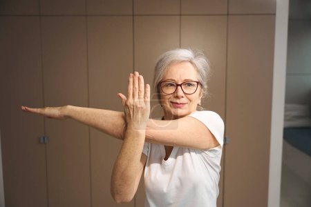 Photo for Pleasant grandmother in glasses is standing in an asana, female is training at home - Royalty Free Image