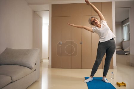 Photo for Joyful elderly woman stands on a karimate in a triangle pose, the female is engaged at home - Royalty Free Image