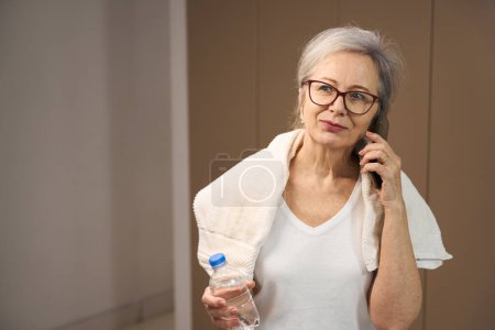Photo for Gray-haired pensioner is talking on the phone during a home workout, a woman has a towel on her shoulders - Royalty Free Image