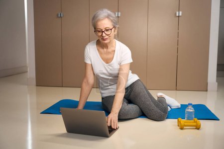 Photo for Elderly lady sits on a carimat in front of a laptop, near water and dumbbells for sports - Royalty Free Image