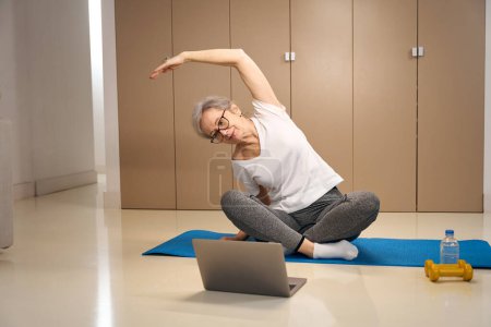 Téléchargez les photos : Older woman does an online workout in front of a laptop, next to a bottle of water and dumbbells - en image libre de droit
