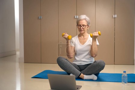 Foto de Mujer mayor se sienta con mancuernas en las manos en un karimate, delante de ella hay un ordenador portátil - Imagen libre de derechos