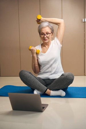 Téléchargez les photos : Pretty elderly pensioner doing exercises with dumbbells in front of a laptop, a woman sits on a carimate - en image libre de droit