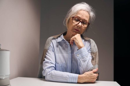 Foto de Triste anciana se sienta sola en una mesa con un chal en los hombros - Imagen libre de derechos