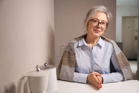 Foto de Mujer bastante mayor se sienta en casa en la mesa de la cocina, tiene un chal en los hombros - Imagen libre de derechos