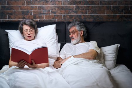Photo for Old couple lying on bed in bedroom, wife reading book to her husband before sleep - Royalty Free Image