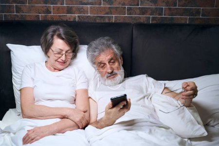Téléchargez les photos : Elderly lady and man lying on bed in bedroom, male holding mobile phone, reading news - en image libre de droit