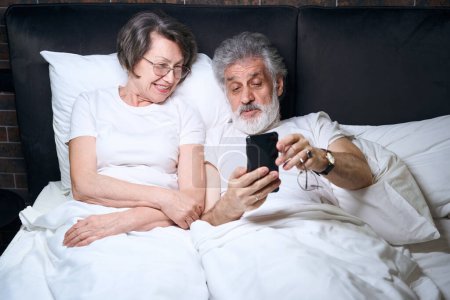 Photo for Elderly lady and man lying on bed in bedroom, male holding mobile phone, having video call - Royalty Free Image
