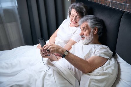 Photo for Elderly couple lying on bed in bedroom, male holding mobile phone, having video call - Royalty Free Image