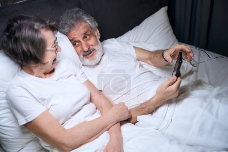 Photo for Elderly woman and male lying on bed in bedroom, male holding mobile phone, looking at wife - Royalty Free Image