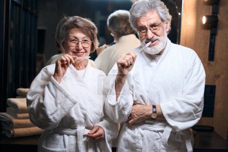 Téléchargez les photos : Femme âgée et homme en manteaux blancs debout dans la salle de bain, tenant une brosse à dents et regardant la caméra - en image libre de droit