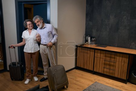 Foto de Elderly woman and man entered rented hotel room, holding bags and looking around the room - Imagen libre de derechos
