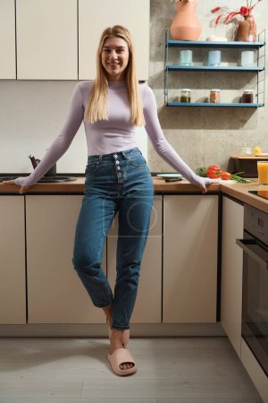 Téléchargez les photos : Portrait grandeur nature d'une jeune femme heureuse souriante appuyée contre le comptoir de la cuisine et regardant devant elle - en image libre de droit
