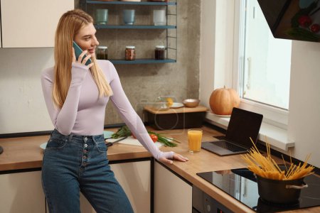 Téléchargez les photos : Smiling young woman leaning on kitchen counter and talking on mobile phone - en image libre de droit