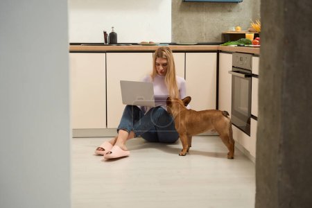 Foto de Mujer grave concentrada sentada en el suelo trabajando en un ordenador portátil en presencia de su mascota - Imagen libre de derechos