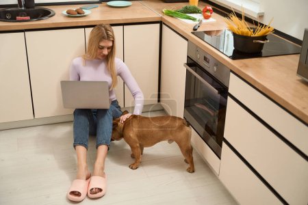 Téléchargez les photos : Young woman with laptop sitting on kitchen floor and stroking her pet - en image libre de droit