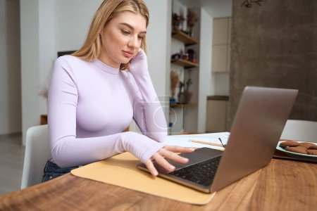 Téléchargez les photos : Calme concentré jeune femme assise à la table de cuisine regardant l'écran d'ordinateur portable - en image libre de droit