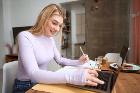 Téléchargez les photos : Femme indépendante souriante avec crayon à la main assise à la table tapant sur un ordinateur portable - en image libre de droit