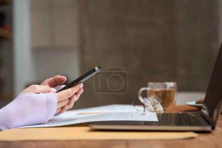 Photo for Cropped photo of female hands holding cellphone in front of portable computer - Royalty Free Image