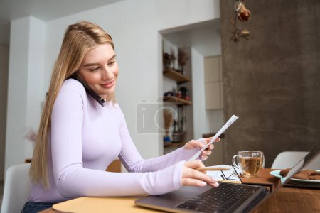 Foto de Mujer joven agradable con hoja de papel en la mano escribiendo en el ordenador portátil durante la conversación telefónica - Imagen libre de derechos