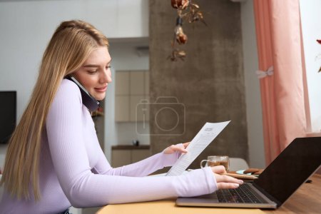 Photo for Pleased young woman with document in hand typing on portable computer and talking on cellphone - Royalty Free Image