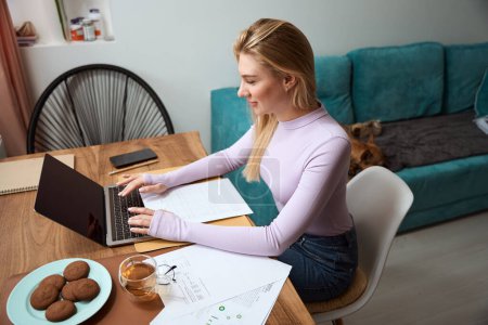 Téléchargez les photos : Side view of smiling female freelancer seated at window table typing on laptop while her dog lying on sofa - en image libre de droit