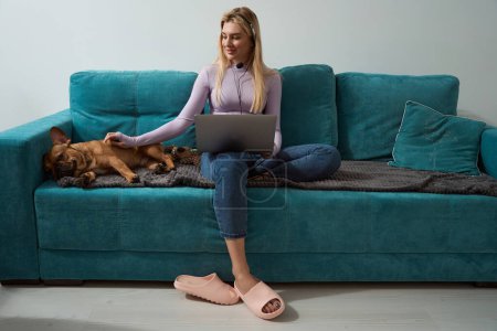 Photo for Pleased young woman with laptop sitting on sofa and stroking her French bulldog - Royalty Free Image