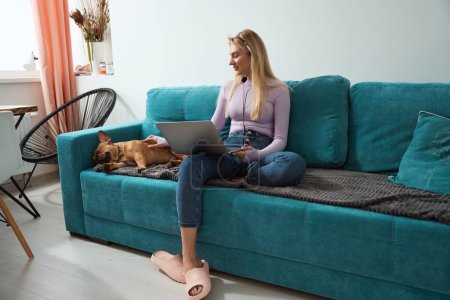 Photo for Calm young female with laptop sitting on sofa and stroking her bulldog - Royalty Free Image