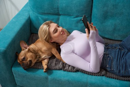 Photo for Smiling young woman with mobile phone in hands lying on her French bulldog - Royalty Free Image