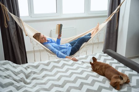 Photo for Joyful female with book in hand lying in hammock and looking at her dog - Royalty Free Image