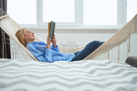 Téléchargez les photos : Vue latérale de la jeune femme détendue avec le livre couché dans l'hamac à l'intérieur - en image libre de droit