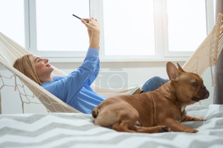 Photo for Smiling contented female taking selfie in hammock while her dog lying on bed - Royalty Free Image