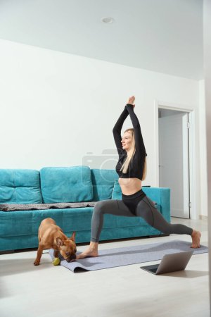 Photo for Joyous fit female doing lunge with arm raise on yoga mat in presence of her pet - Royalty Free Image