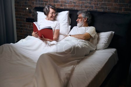 Foto de Anciana y hombre acostados en la cama en el dormitorio, esposa leyendo libro a su marido antes de dormir - Imagen libre de derechos