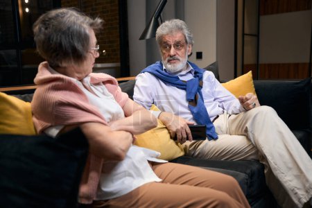 Téléchargez les photos : Homme barbu aux cheveux gris et femme âgée arranger les choses, le couple s'est installé sur un canapé dans une chambre d'hôtel - en image libre de droit