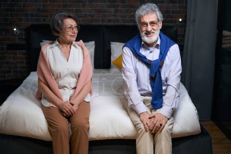 Téléchargez les photos : Heureux homme aux cheveux gris et son compagnon sont assis sur un grand lit, le couple séjourne dans un hôtel confortable - en image libre de droit