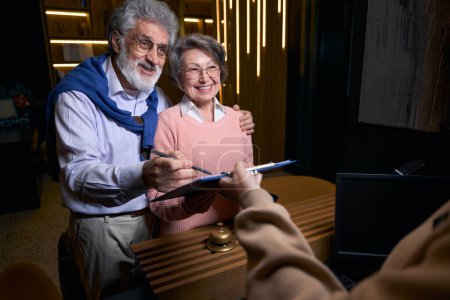 Photo for Hotel staff registers elderly spouses at the reception desk, pensioners in travel clothes - Royalty Free Image