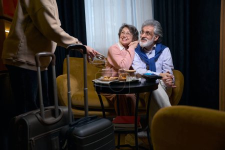Foto de Los jubilados viajeros sonrientes se registran en el hotel, el personal los invita al té - Imagen libre de derechos