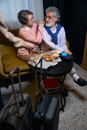 Photo for Traveling elderly couple resting from the road in the hotel cafe, the staff treats them to tea - Royalty Free Image