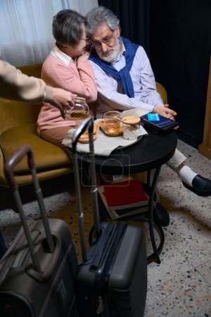 Foto de Abuelos modernos están descansando de la carretera en un café del hotel, el personal los invita a tomar el té - Imagen libre de derechos