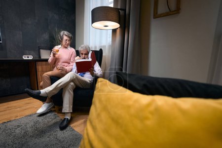 Photo for Elderly couple comfortably settled in an armchair by the window with a book and drinks, people in casual clothes - Royalty Free Image