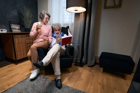 Téléchargez les photos : Homme âgé assis dans un fauteuil près de la fenêtre avec un livre et à boire, sa femme s'assit à côté de lui - en image libre de droit