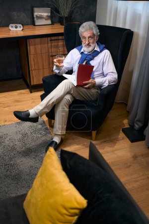 Foto de Hombre mayor se sienta en sillón junto a la ventana con un libro y bebida, tiene el pelo gris y una barba - Imagen libre de derechos