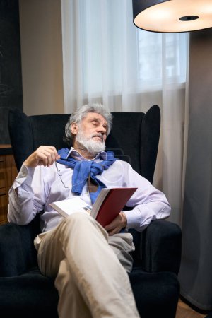Téléchargez les photos : Le vieil homme fait une sieste dans un fauteuil confortable avec un livre, il s'est endormi en lisant - en image libre de droit