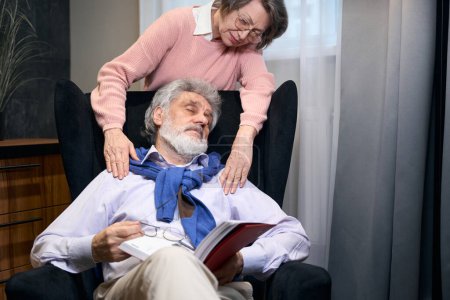Téléchargez les photos : Grand-père barbu s'est endormi dans un fauteuil confortable avec un livre, une dame âgée l'étreint par les épaules - en image libre de droit