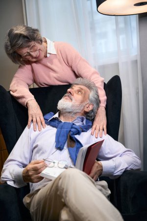Foto de Abuelo barbudo se estableció en un sillón acogedor con un libro, una anciana lo abraza por los hombros - Imagen libre de derechos